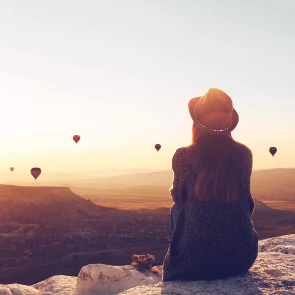 View from the back of a girl in a hat sits on a hill and looks at air balloons in Cappadocia in Turkey.